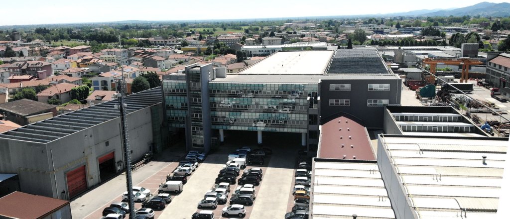 Utsikt över en industriell anläggning med flera byggnader och parkerade bilar, omgiven av en tät bostadsområde och gröna fält i bakgrunden.