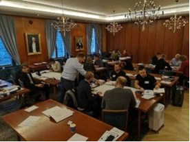 Ett klassrum med deltagare som arbetar vid bord, där en instruktör hjälper en grupp. Rummet har träpaneler och stora fönster med draperier.