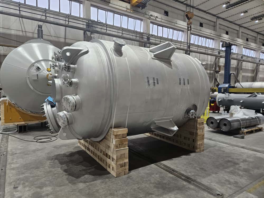 En stor tryckkärl i aluminium med flera anslutningar och detaljer, placerad på en pall i en fabrikshall. I bakgrunden syns ytterligare cylindriska behållare och industriell utrustning.