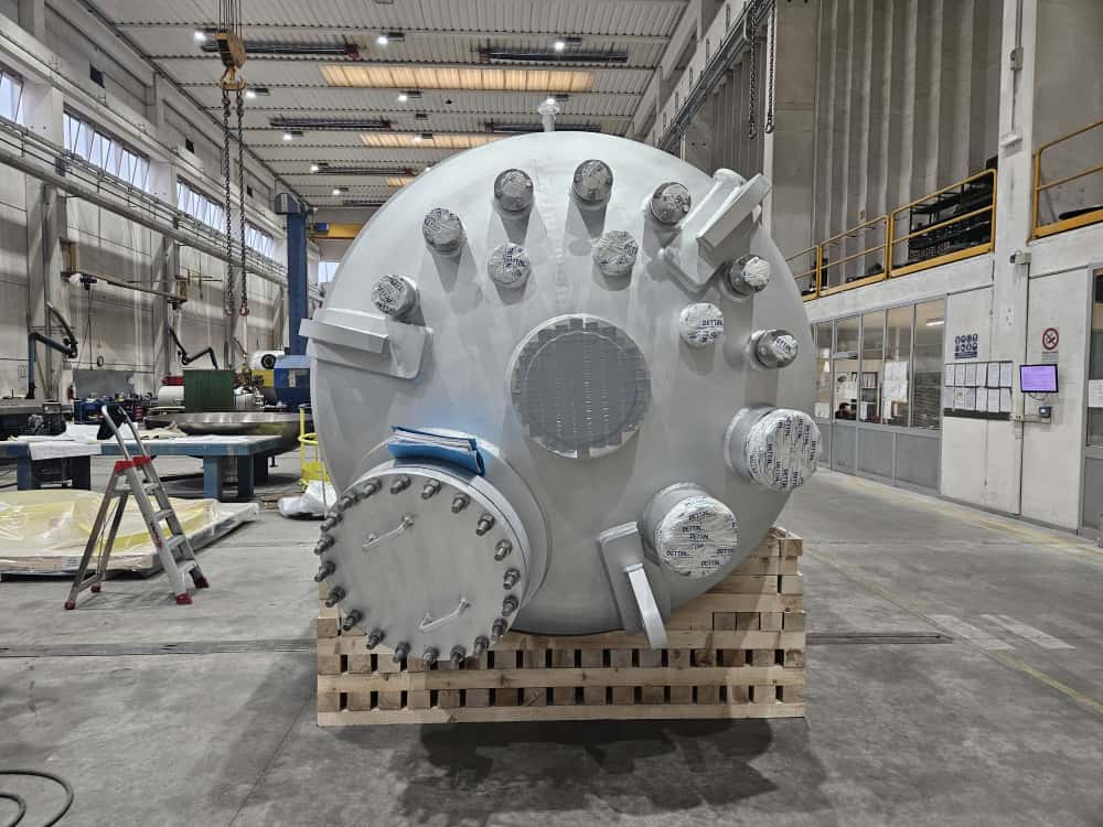 En stor aluminiumtryckkärl med flera anslutningar, ligger på en pall i en fabriksmiljö, omgiven av industriell utrustning.