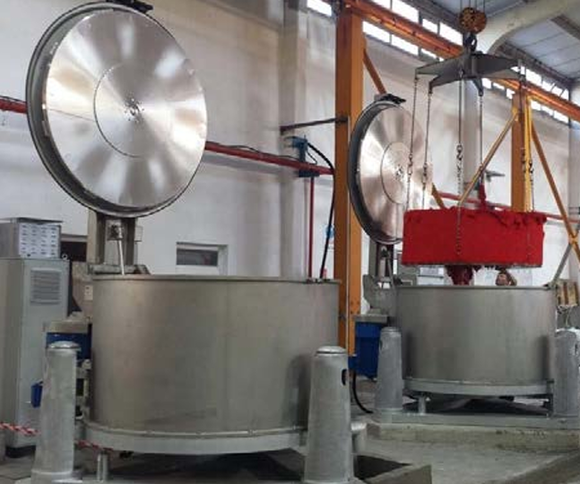 Två stora, metalliska textilmaskiner står i en fabrik med silvriga lock och en kran som lyfter en röd behållare.