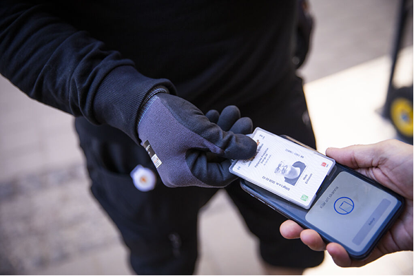 En person som visar sitt ID06-kort för en mobil enhet, med en hand som håller kortet och en annan hand som sträcker sig mot mobilen.