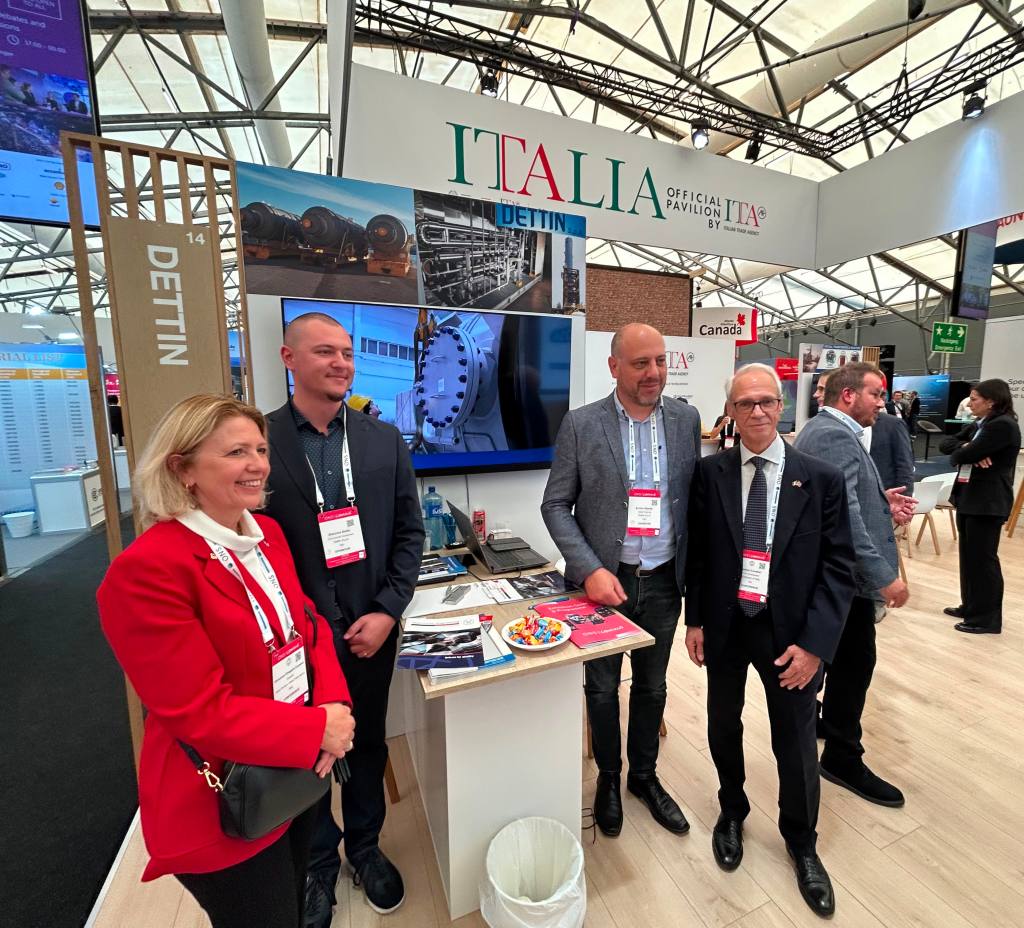 Fyra personer står framför en monter som representerar Italien, med skylten 'ITALIA' och 'DETTIN'.